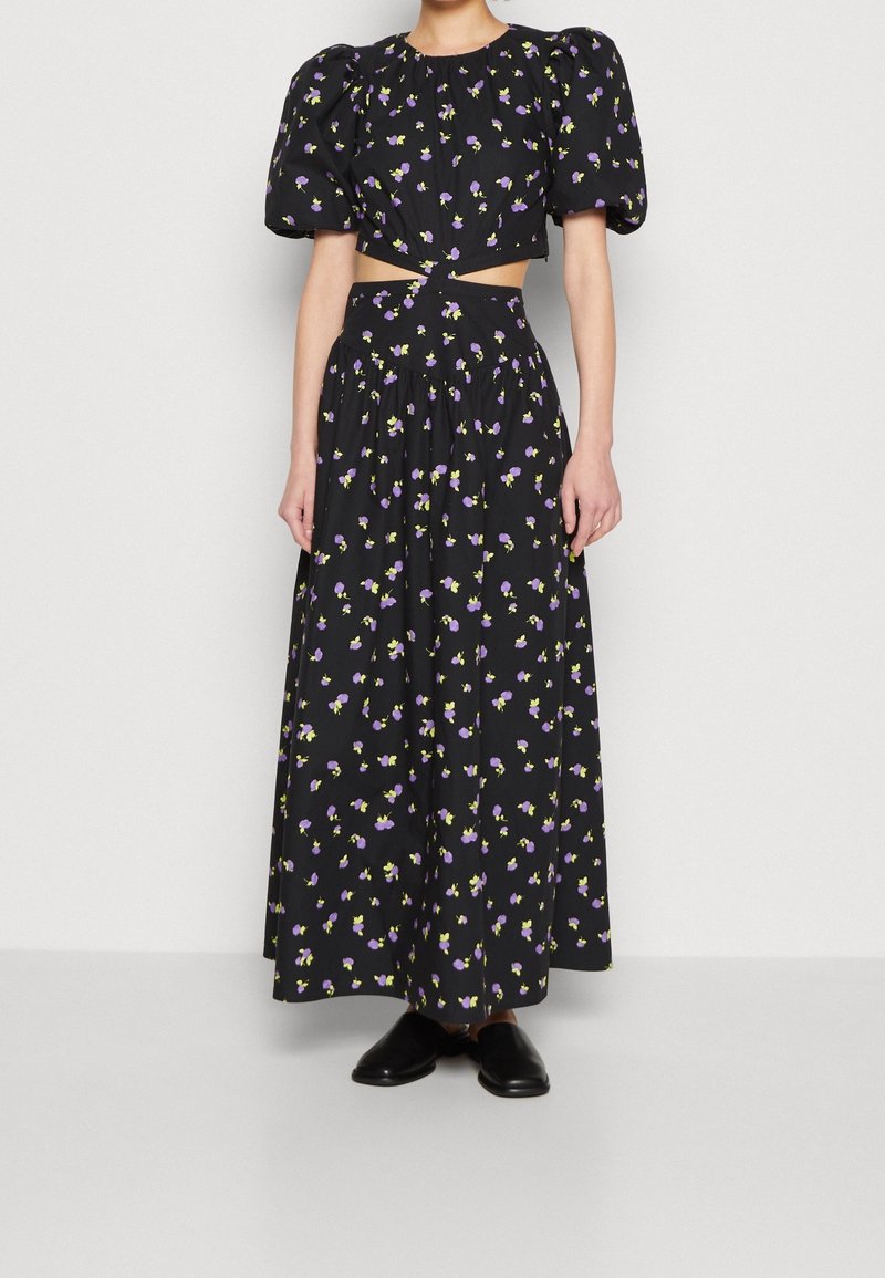 Femme portant une robe noire fleurie deux pièces avec des manches courtes bouffantes, une taille découpée, une longue jupe et des chaussures noires sans lacets.