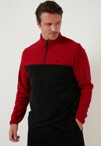 Crveni i crni flis pulover s polu-zip zatvaranjem, s crnim logotipom na prsima i kontrastnim gornjim i donjim blokovima boja.