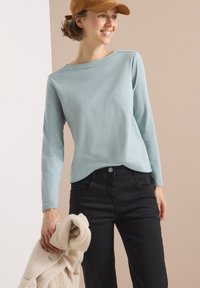 Lichtblauw langemouwen shirt met een losse pasvorm, gecombineerd met zwarte broek; houdt een beige, textuur, fluffy jack vast. Een bruine pet maakt de outfit compleet.