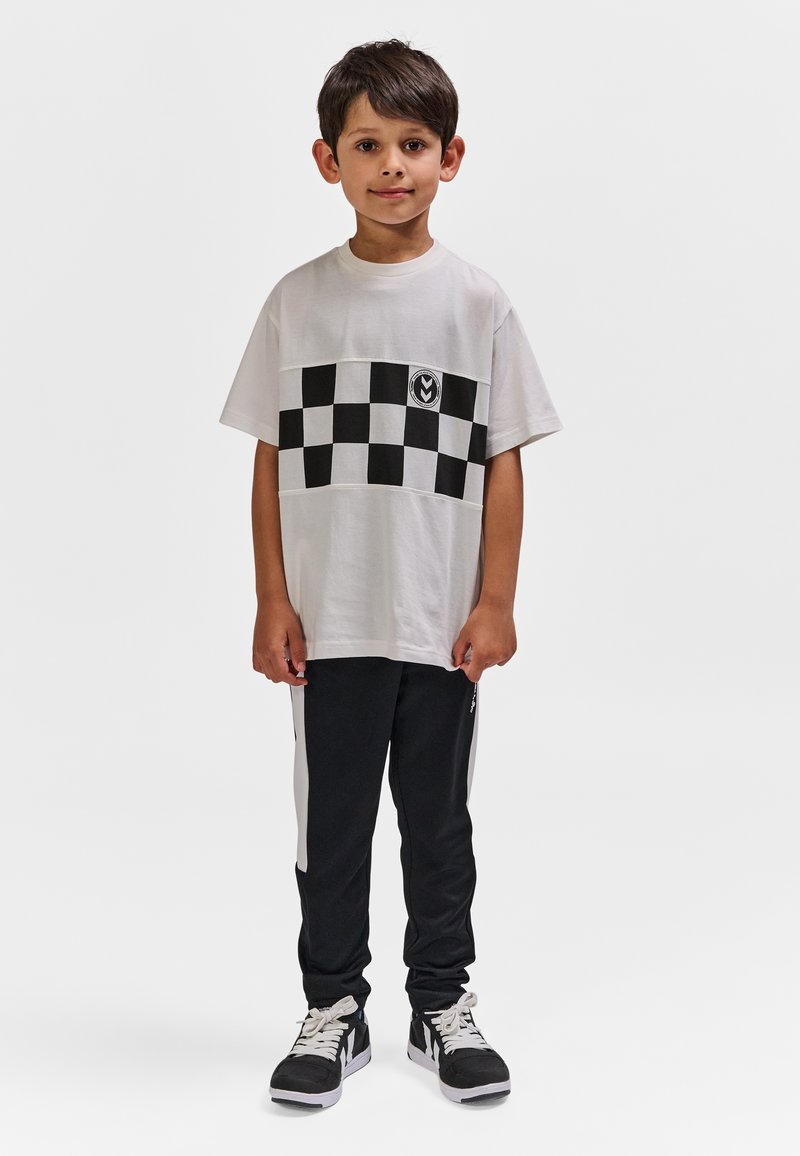 Young boy standing wearing a white T-shirt with black checkerboard pattern and black track pants with white side stripes and black sneakers.