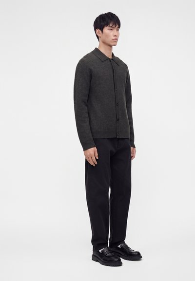 Homme debout portant un cardigan boutonné gris foncé, un pantalon noir et des mocassins noirs sur un fond clair uni.