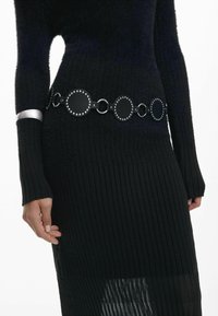 Cintura in pelle nera con anelli circolari e borchie. Indossata sopra un capo nero a coste, con maniche lunghe e un design dell'orlo a strati.