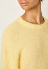 Rumena, mehka pulover v ohlapnem dizajnu z okroglo izrezano obleko in rahlo puhasto teksturo. Osredotočite se na predel vratu in ramen.