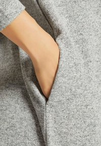 Grauer Stoff mit einer weichen Textur, ausgestattet mit einer seitlichen Tasche, die von einer Hand erreicht wird. Das Material hat ein dezentes meliertes Muster.