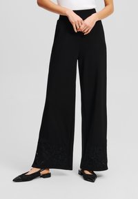Zwarte wijde broek van gladde stof, met bloemenborduursel aan de zoom en een hoge tailleband. Gepaard met zwarte platte schoenen.