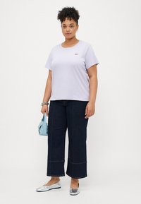 Camiseta corta de manga en color lila con un pequeño logo rojo, combinada con pantalones de mezclilla anchos en color oscuro. Los accesorios incluyen un bolso azul y zapatos planos plateados.