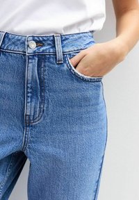 Blauwe denim jeans met hoge taille, vervaagde details, contrasterend stiksel en een voorzak, gedragen met een wit shirt.