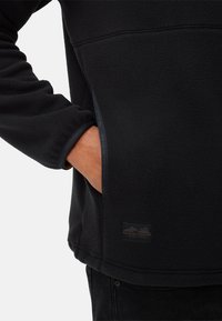 Zwarte fleece pullover met een gestructureerd oppervlak, zijzakken en een label met bergafbeeldingen en tekst aan de zoom.