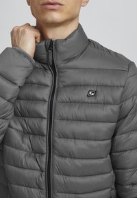 Mees tõmbab lukku halli tikitud jope, millel on kõrge krae ja väike must-valge logo rinnal.