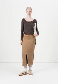 Junge Frau steht und hält eine weiße Handtasche, trägt ein braunes Langarmoberteil, einen beigen Midirock mit Schlitz vorne und Sneakers mit Leopardenmuster.