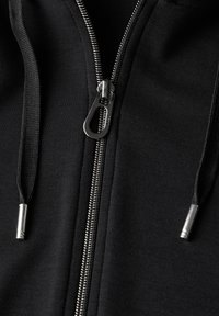 Tissu de sweat-shirt noir avec une fermeture éclair en argent, présentant des cordons avec des embouts métalliques et une texture lisse. Gros plan sur le détail de la fermeture éclair.