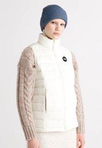 Witte gewatteerde vest met hoge kraag, rits aan de voorkant en rond logo. Model draagt een blauwe geribbelde breimuts en beige kabelgebreide lange mouwen.