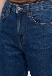 Gros plan sur un jean en denim bleu avec des coutures orange, un bouton en métal et des passants de ceinture porté avec un haut marron.
