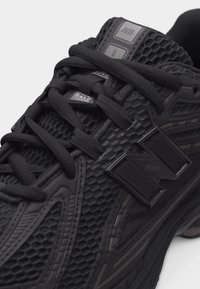 Gros plan sur une chaussure de course noire New Balance avec une tige en mesh texturé, des lacets noirs et un logo "N" en relief sur le côté.