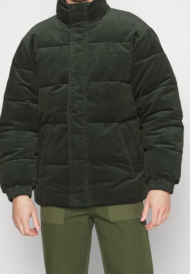 Chaqueta de plumas de pana verde con cuello alto, puños elásticos y bolsillos frontales. Textura suave con diseño acolchado.