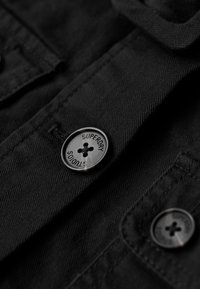 Camisa de tela negra que presenta un primer plano detallado de botones redondos negros grabados con "SUPERDRY" y un tejido texturizado.