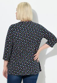 Frau mit kurzen blonden Haaren, die ein marineblaues Oberteil mit Mustern von Teddybären, einem Fußball und pinken Helmen sowie Jeans trägt und mit der Hand in die Hüfte posiert.