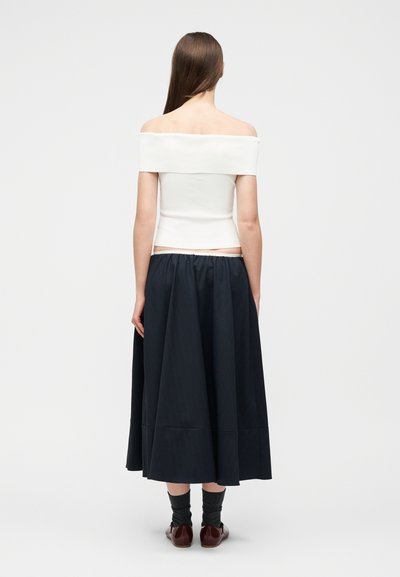 Γυναίκα με μακριά καστανά μαλλιά, γυρισμένη προς τα πίσω, φοράει λευκό τοπ με ακάλυπτους ώμους, μπλε midi φούστα, σκούρες κάλτσες και καφέ παπούτσια.