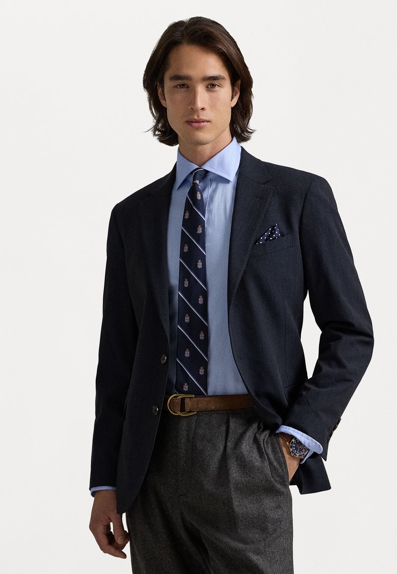 Blazer bleu marine foncé sur une chemise bleu clair, avec une cravate à motifs bleus et beiges. Un pantalon gris et une ceinture marron complètent le look.