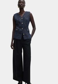 Marineblauw tweedvest met V-hals, voorzien van gouden knopen en steekzakken aan de voorkant, gecombineerd met zwarte broek met wijde pijpen. Soepele textuur, perfect op maat gemaakt.