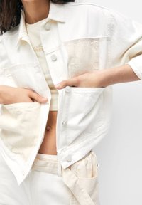 Biała dżinsowa kurtka charakteryzuje się luźnym krojem, srebrnymi zatrzaskami i kontrastowymi teksturowanymi panelami. W połączeniu z dopasowaną białą bluzką i spodniami.