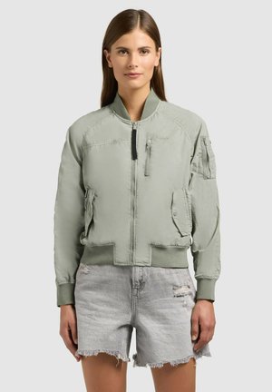 Žena v svetlo zelenej bomber bunde s rebrovanými manžetami a lemom, v kombinácii so svetlo sivými roztrhanými riflovými šortkami.