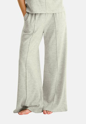 Personne portant un pantalon large gris clair avec des coutures à l'avant, les mains dans les poches, debout pieds nus sur un fond blanc.