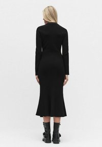 Musta pitkähihainen mekko, jossa on vartalonmyötäinen siluetti, liehuva helma, ribattu pinta, ja joka on yhdistetty mustiin nilkkureihin.