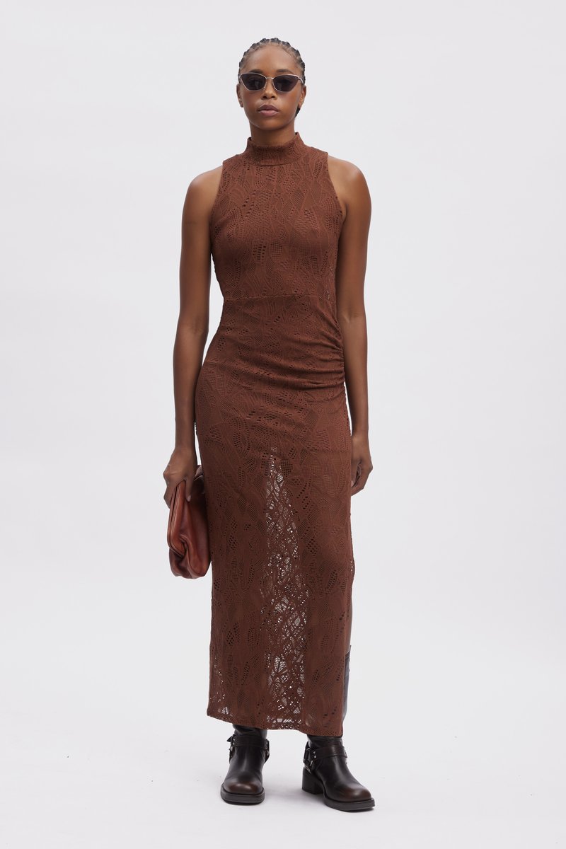 Brązowa maksiszata z koronką, z wysokim dekoltem i marszczeniami po bokach, w połączeniu z czarnymi botkami za kostkę i brązowym kopertówką, na jednolitym tle.