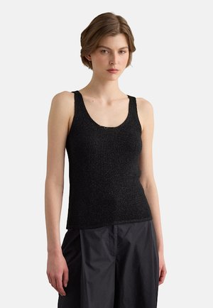 Jonge vrouw met kort bruin haar, die een mouwloze zwarte getextureerde tanktop en losse zwarte broek draagt, staand tegen een eenvoudige lichte achtergrond.