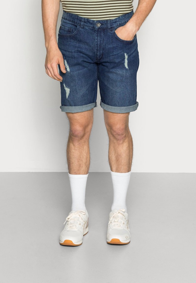 Shorts en denim bleu foncé avec des accents usés et des revers, assortis à des chaussettes blanches et des baskets beige et orange.