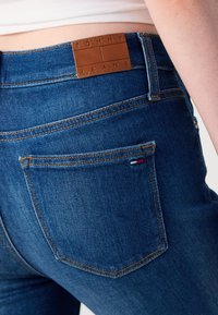 Sötétkék, farmer anyagú nadrág, barna bőr címkével, amelyen a "TOMMY JEANS" felirat található a derékrészen, látható varrással és egy kis logóval a hátsó zseben.