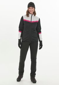 Schwarze und weiße Sportjacke mit pinken Akzenten, Reißverschluss vorne und Taschen, kombiniert mit schwarzen Hosen und Handschuhen, getragen von einer lächelnden Person.
