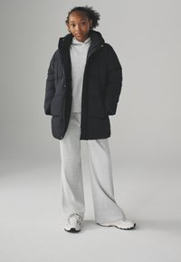 Sort pufjakke med høj krave, parret med en lys grå ribbet sweater og brede bukser. Hvide sneakers med sorte såler.