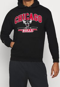 Sudadera negra con el logo de los Chicago Bulls en rojo y blanco, con capucha con cordón y bolsillo tipo canguro en la parte frontal. Textura de tela suave.