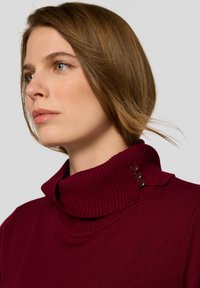 Bordeauxfarbener Rollkragenpullover mit gerippter Struktur und drei Knöpfen am Hals. Nahaufnahme der Seitenansicht mit glatten Stoffdetails.