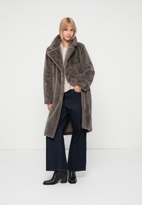 Palton din blană artificială gri, cu un guler lat și mâneci lungi, având un design cu deschidere frontală. Combinate cu blugi negri cu croială largă și cizme negre cu toc.