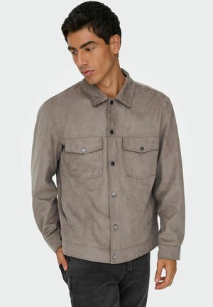 Un jeune homme portant une veste en daim brun clair avec des boutons-pression à l'avant et des poches poitrine, regardant à sa gauche les mains dans les poches.