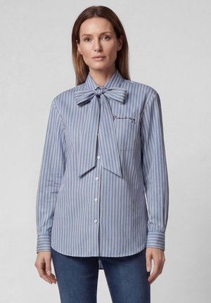 Woman wearing a blue and white striped button-up shirt with a large bow tie at the neck and embroidered text on the chest.