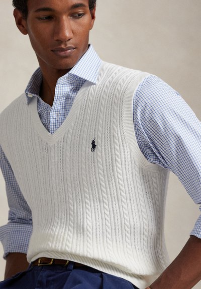 Λευκό πλεκτό, αμάνικο πουλόβερ με ανάγλυφη πλέξη πάνω από μπλε καρό πουκάμισο· διαθέτει λαιμόκοψη σε σχήμα V και μικρό κεντημένο λογότυπο στο στήθος.