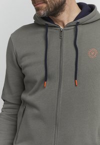 Grey zip-up hoodie with navy accents, orange-tipped drawstrings, a small logo on the chest, and side pockets. Soft fabric texture.
