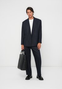 Young man wearing a dark pinstripe suit and white shirt, holding a black leather tote bag, standing against a plain white background.