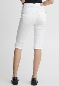 Pantalons capri en denim blanc avec une coupe ajustée, revers aux chevilles, poches arrière et accents de boutons noirs. Portés avec des chaussures noires.