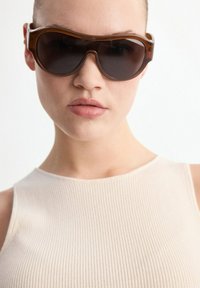 Jeune personne aux cheveux courts portant de grandes lunettes de soleil marron et un haut beige sans manches à côtes, sur un fond uni.