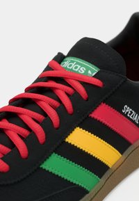 Μαύρα παπούτσια Adidas Spezial με κόκκινες κορδόνες, πράσινες, κίτρινες και κόκκινες λωρίδες, πράσινη ετικέτα λογότυπου και σόλα από καουτσούκ.