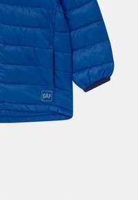 Doudoune bleue en polyester brillant, avec des sections matelassées, des poignets côtelés et un petit logo GAP sur l'ourlet inférieur.