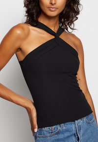 Mujer vestida con un top negro sin mangas y con un escote retorcido, combinado con jeans de mezclilla azules, posando con una mano en la cadera.