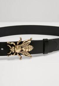 Black leather belt featuring a prominent gold bee-shaped buckle with detailed wings and body texture. Adjustable fit with a smooth finish.