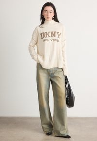Maglione lavorato a maglia color crema con la scritta "DKNY NEW YORK", abbinato a jeans ampi blu chiaro e una borsa nera, su uno sfondo neutro.
