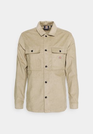 Beige corduroy langærmet skjortejakke med knapper foran, to knappede brystlommer og et lille logopatch på venstre lomme.